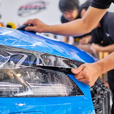 Close up of a technician applying clear paint protection film to the front of a blue car, smoothing the film over the headlight and hood. The image supports the section about protecting vehicle paint from chips, scratches, and road debris with a precise installation.