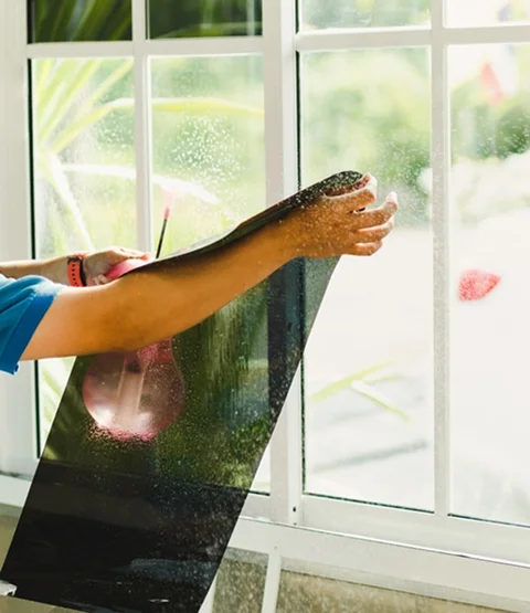 Technician applying dark window film to a residential window from the inside while smoothing it across the glass. The image supports the section about professional installation of security and safety film to strengthen windows and reduce breakage risks.