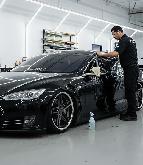 A technician installs window tint on a black car inside a clean shop, stretching film across the side window during the application process. The image supports the section about automotive tint for heat reduction, UV protection, and a clean factory style finish.